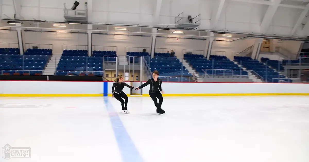 A young adult female figure skater in a black synthetic outfit practicing with her male counterpart on an indoor ice rink, surrounded by empty spectator seats under artificial lighting.