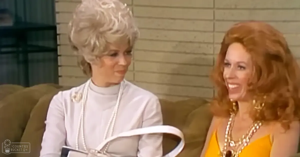 Middle-aged female in white turtleneck sweater with pearl necklace, holding book or notebook, seated on green sofa. Another middle-aged to older adult female in yellow sleeveless top and layered gold necklaces nearby. Indoor domestic setting.