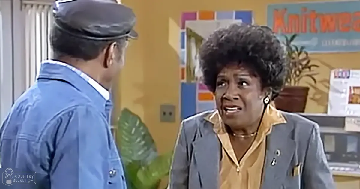 A middle-aged woman in a light gray blazer and gold blouse stands indoors with a man wearing a blue denim jacket and white shirt, surrounded by indoor decor including plants and a 'Knitwea' sign.