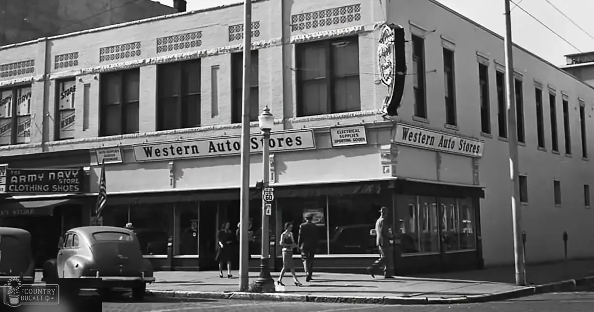 Urban street corner with 'Western Auto Stores' signage in an urban area; vintage car parked on the curb, lamppost, and various product signs indicating electrical supplies, sporting goods, and clothing are visible.