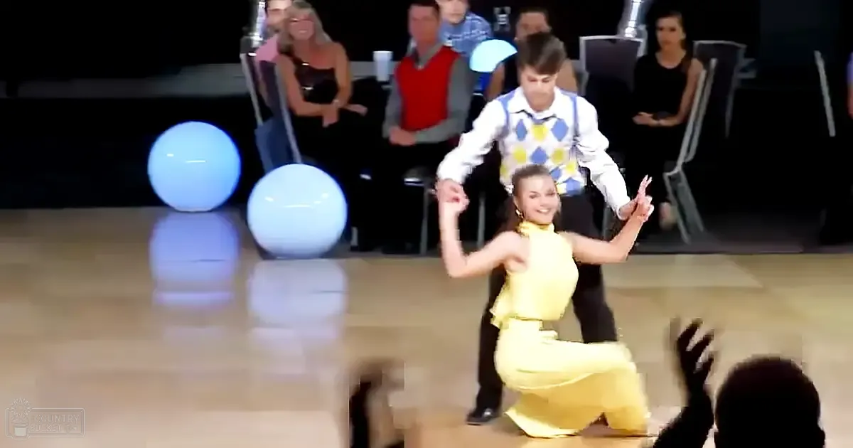 Young adult dancers perform at an indoor ballroom competition, with one wearing a yellow satin dress and another in a white shirt with blue diamond patterns.