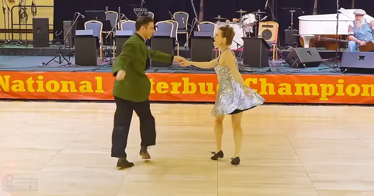 A dance performance at an indoor event, with a man in a green blazer and a woman in a silver sequined dress. The setting includes a stage setup with musical instruments and a bright orange banner reading "National Interbus Championship."