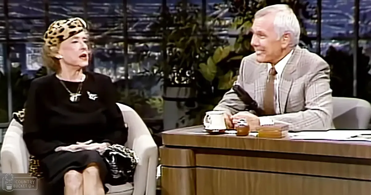 An older adult woman wearing a black dress with a patterned skirt and leopard print headscarf, seated on an upholstered chair in front of a man in a plaid suit jacket. The setting appears to be a studio for a talk show or interview.