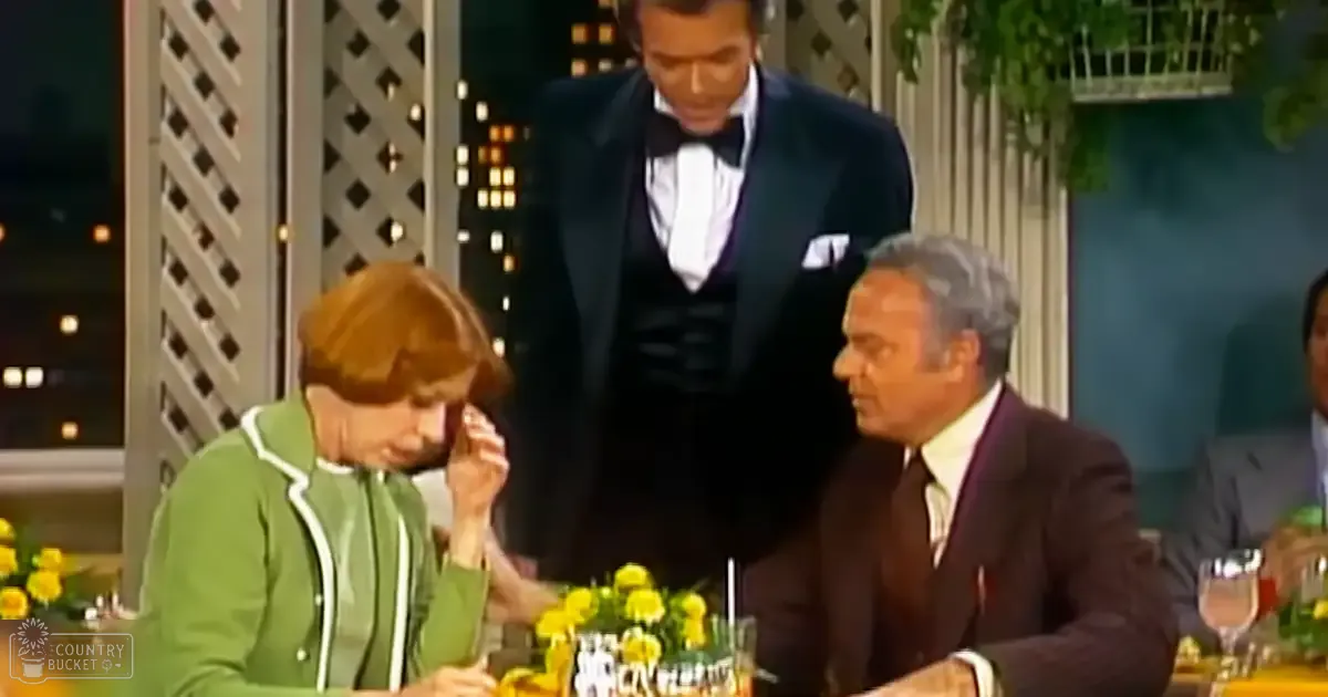 An older adult female wearing a green jacket with white trim, seated at an upscale restaurant or event with yellow flowers on her table and holding something small in her hands; alongside are other guests dressed formally.