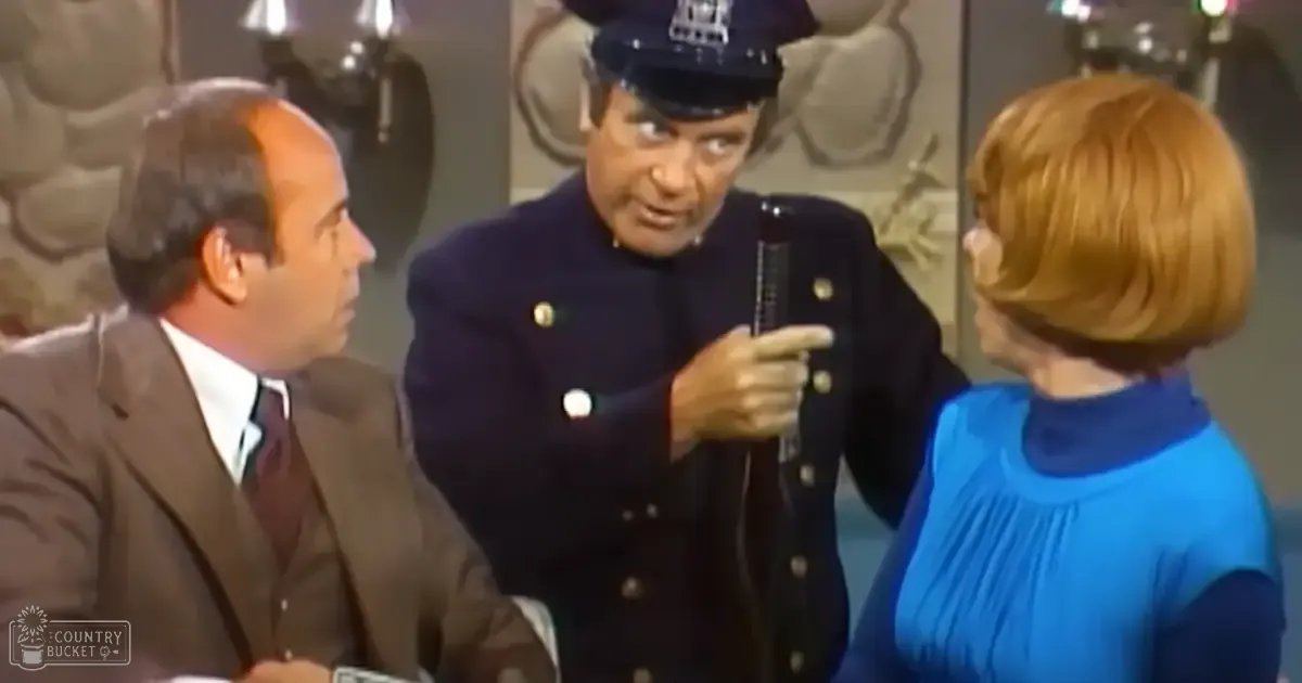 An adult male police officer wearing a dark brown suit jacket, an older adult in a dark blue uniform with gold buttons and peaked cap holding something, and a middle-aged woman in a bright blue sleeveless top or dress. The scene is set indoors, possibly on a television show set.