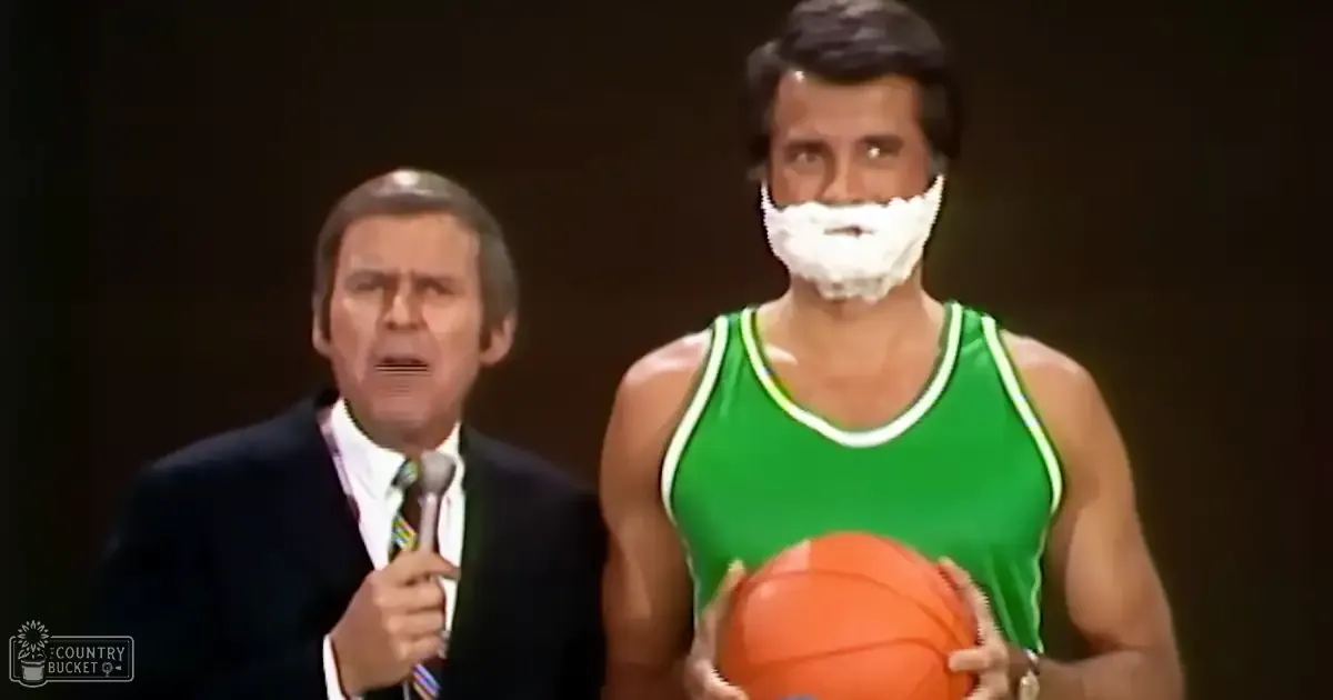 A middle-aged man in a dark suit jacket speaks into a microphone while an older male in a green basketball jersey holds the ball, suggesting an indoor event or performance.