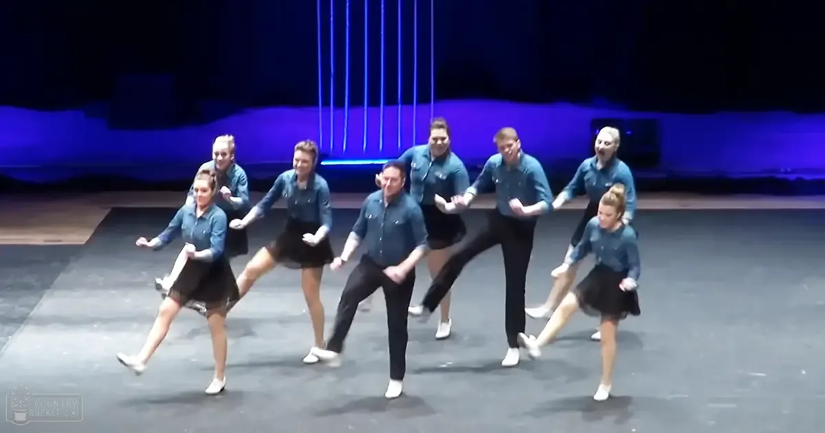 Young adults performing on stage with dark curtains as a backdrop under dim, focused lighting in what appears to be an indoor performance venue.