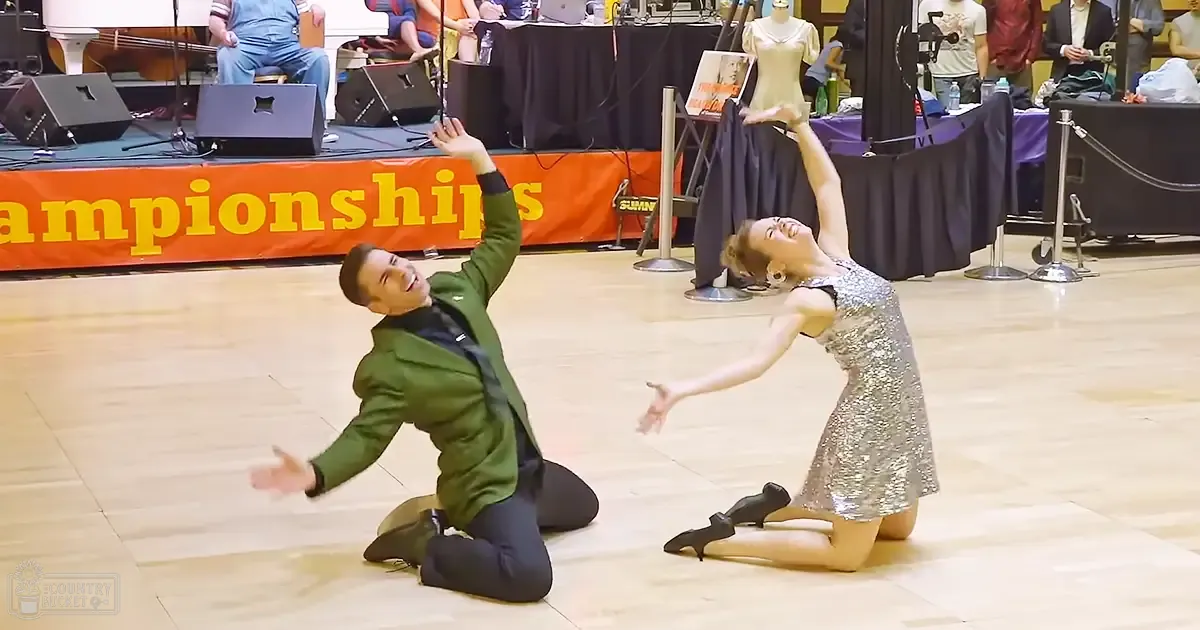 Two young adult dancers performing at 'Championships' event; male in green jacket and black pants, female in silver sequined dress with earrings.