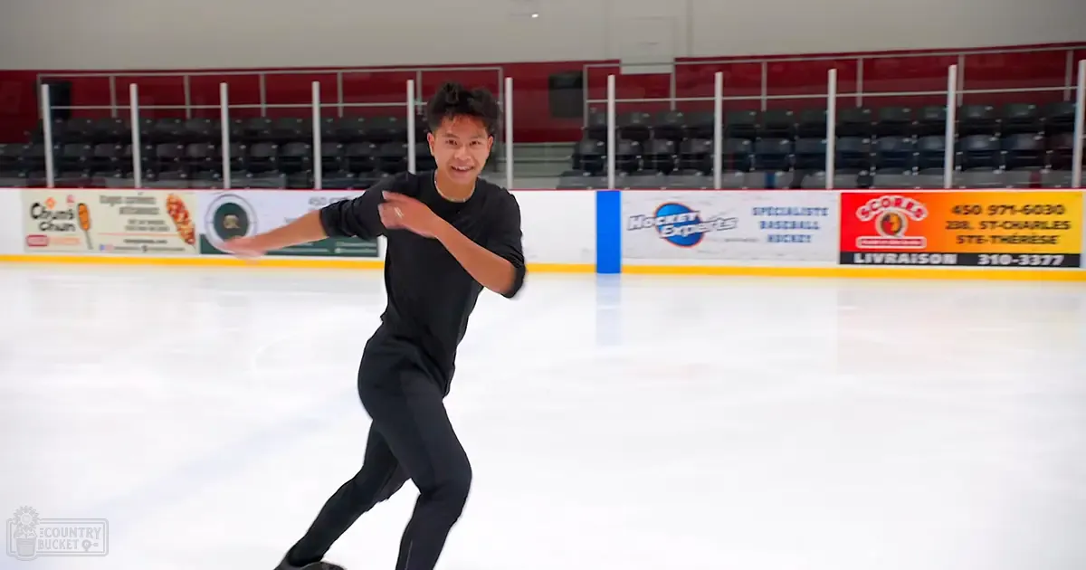 A young adult male wearing a black long-sleeved shirt skates indoors on an ice rink with advertisements visible in the background.