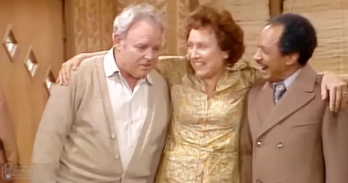 Three individuals in an indoor setting, likely from a television show or sitcom; middle-aged man (left) wearing a light beige cardigan over a white collared shirt, older adult woman (center) with curly brown hair in a gold patterned dress, and another middle-aged man (right) in a light brown double-breasted suit jacket.