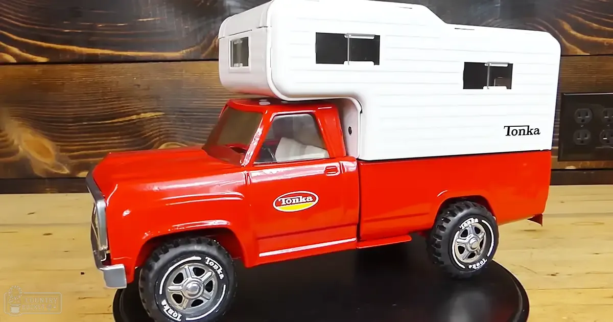 A toy Tonka truck with a white camper trailer on its bed, set against a wooden wall indoors.