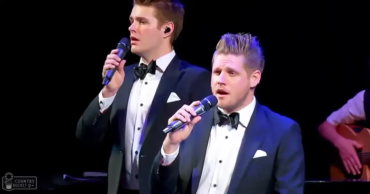 Musical performance with two young adult male singers in dark suits and bow ties against a stage backdrop under dim lighting.
