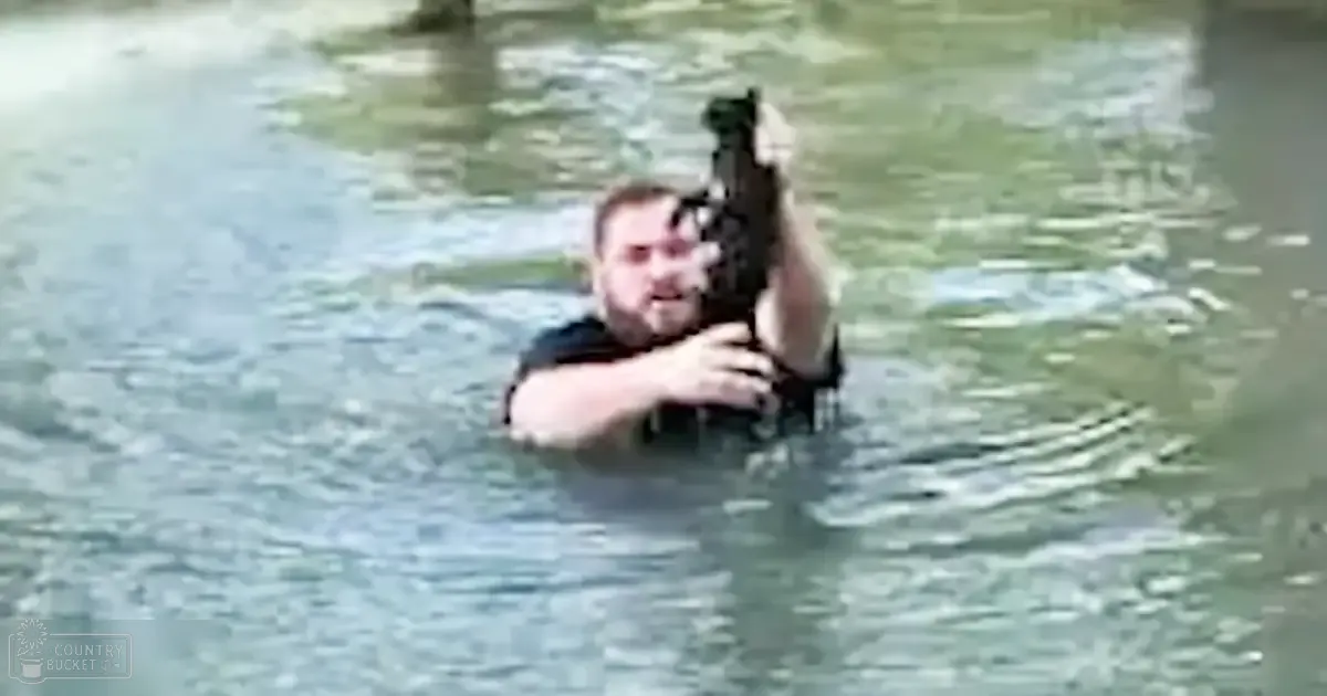 An adult male holding a rifle in an outdoor body of water surrounded by greenery, with reflective daylight.