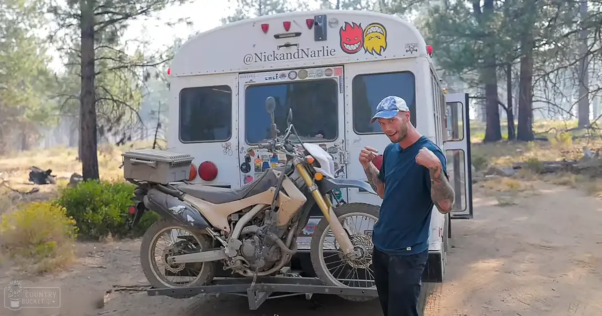 Adult male in blue t-shirt, dark pants, light cap, tattoos, holding small object outdoors near white bus with stickers and cartoon faces.