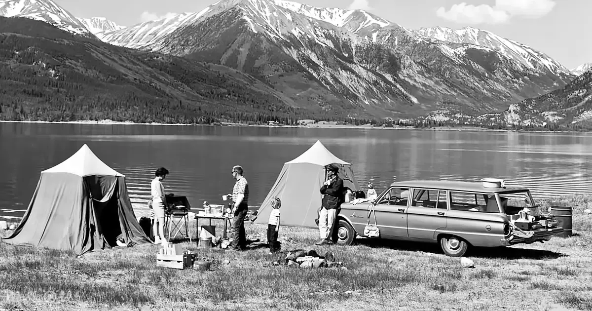 Family camping scene near a lake, surrounded by snow-covered mountains; adults wearing casual outdoor attire, child in dark pants and possibly a light top; vintage station wagon with trunk open, two tents set up, small table with items on it; late spring or summer day with clear skies.
