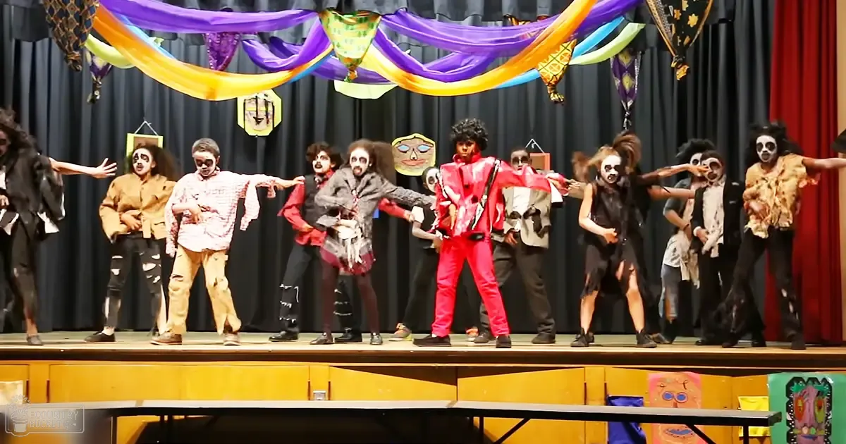 Children and young adults performing in a school auditorium with colorful costumes inspired by zombies, clowns/jesters, and characters from the 1920s-80s.