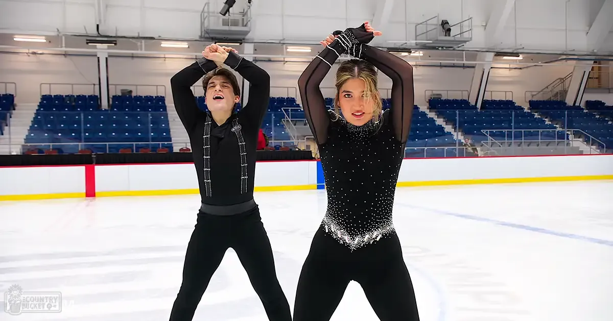 Two young adult figure skaters in an indoor ice rink, surrounded by empty blue spectator seats with railings and lighting fixtures, wearing black and silver outfits typical of skating attire.