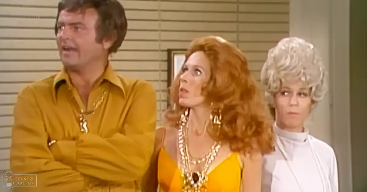An adult male in a relaxed yellow shirt indoors, surrounded by two adult females wearing form-fitting yellow dresses and white turtlenecks, near walls with horizontal blinds and a framed picture or mirror.