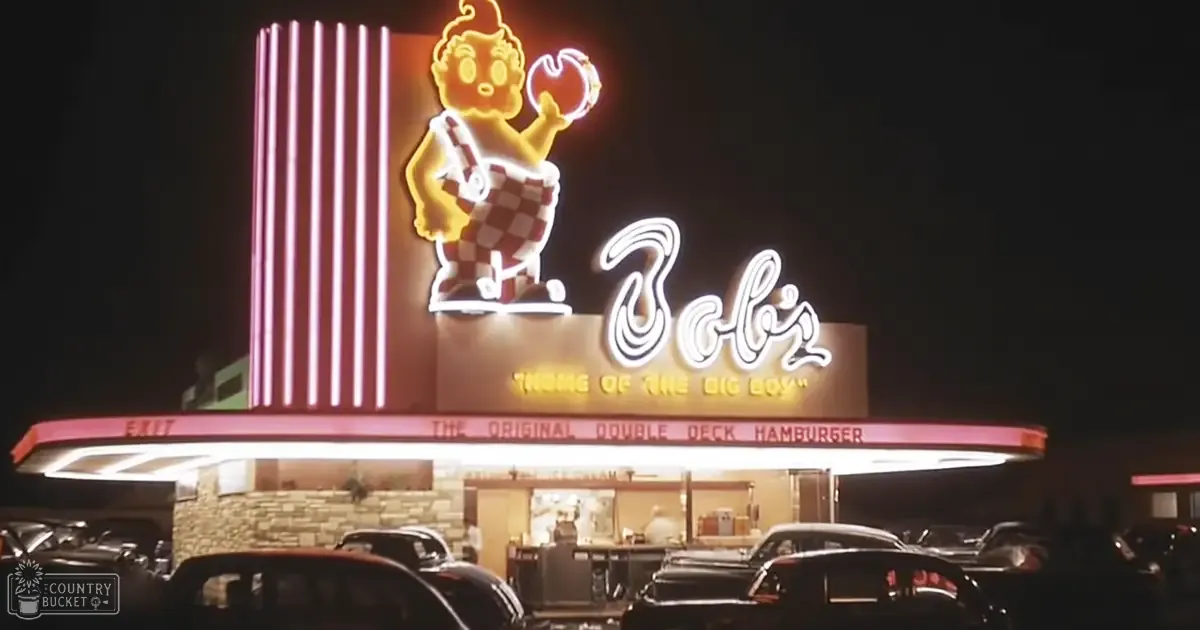 Bob's Big Boy restaurant sign at night, illuminated neon lights with a cartoon character holding an apple, set in a parking lot surrounded by cars.