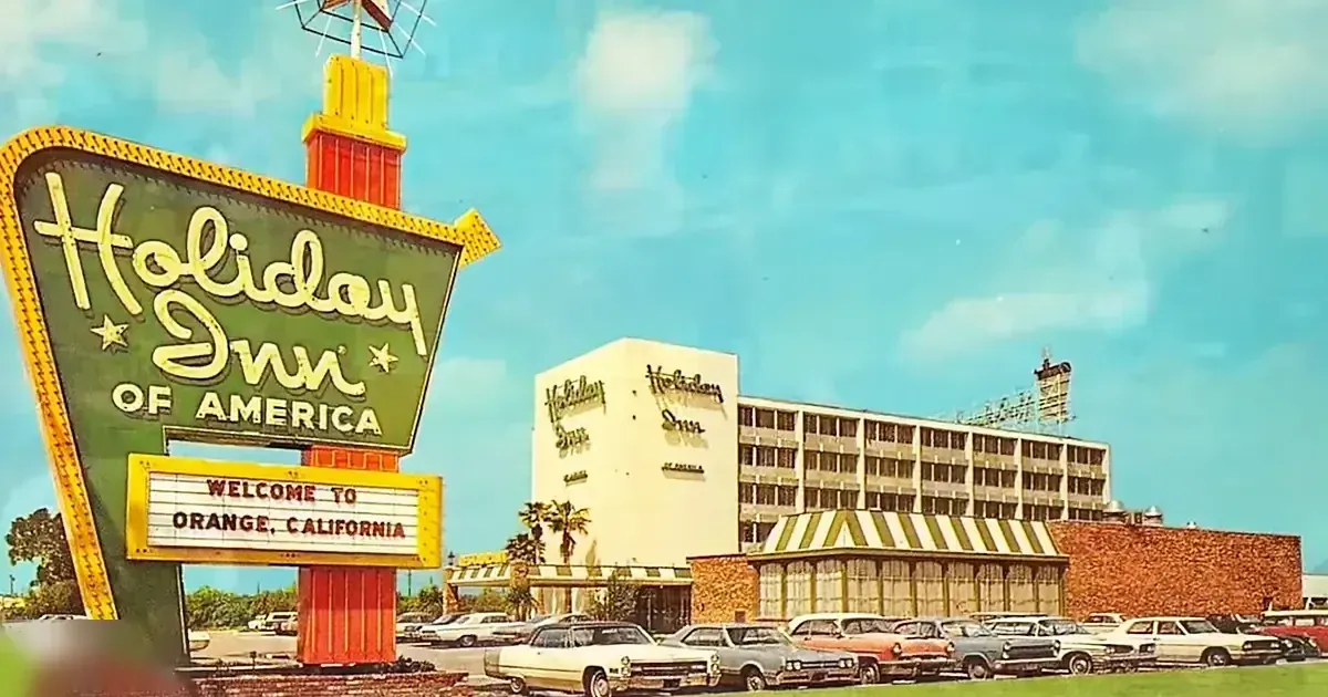 Holiday Inn of America sign on clear day with parked mid-20th century cars and palm trees, Orange, California