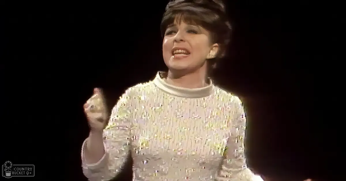 A middle-aged female performer in a 1960s or early 1970s-style sequined top on stage with a microphone, set against a dark background.