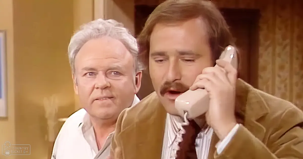 A middle-aged man in a brown jacket and white striped shirt holds an older model phone to his ear while another person stands behind him indoors with beige walls.