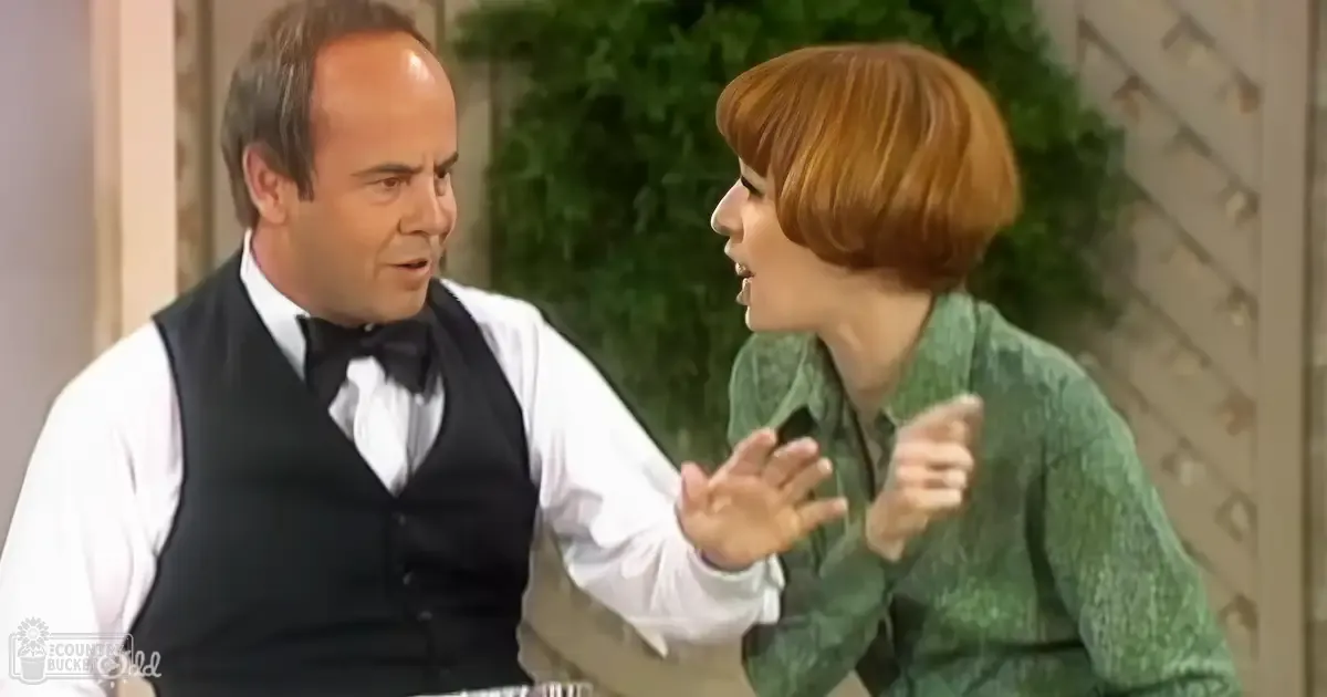 An indoor scene from a television show or film, with two adults dressed formally and informally against greenery with decorative wall elements.