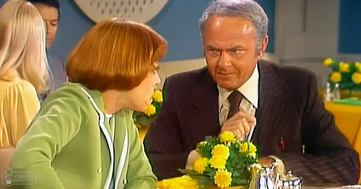 A middle-aged man in a dark pinstriped suit over a white shirt and tie sits at a table with yellow flowers and silver teapots, while a woman in a fitted green cardigan with white trim stands nearby against a blue wall.