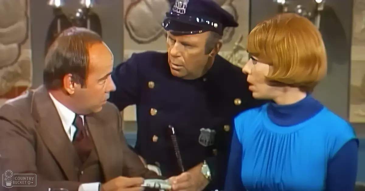 A mid-20th-century television scene set indoors within an office or police station environment with a man in a dark brown suit and white shirt, another middle-aged man in a police uniform with gold buttons and insignia, and a woman wearing a blue sleeveless top. The background includes framed artwork or decorations on the walls.