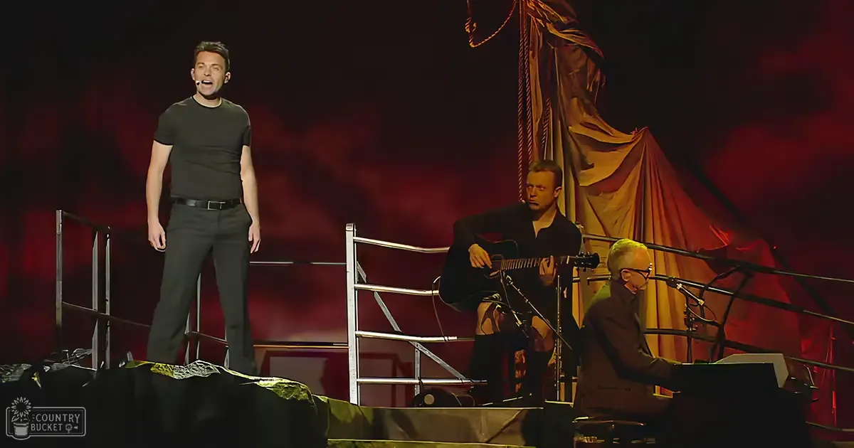 An adult male singer on stage, accompanied by another adult male guitarist and an adult male keyboardist in a dimly lit performance setting with red stage lighting.