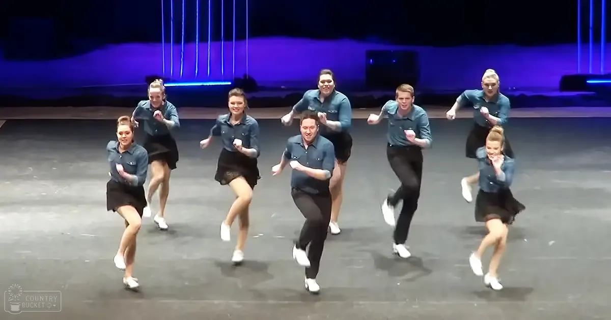 A dance group performing on a lit indoor stage with vertical light beams in the background.