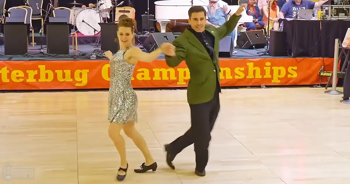 A dance couple performing at an indoor event called 'terbug Championships'. The female dancer wears a silver sequined dress with black shoes, while the male dancer is in a dark green blazer and black trousers. The background shows stage equipment and audience seating.