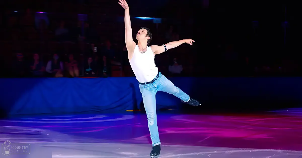 An indoor arena with spectators seated, a young adult male figure skater performing on an ice rink at night.