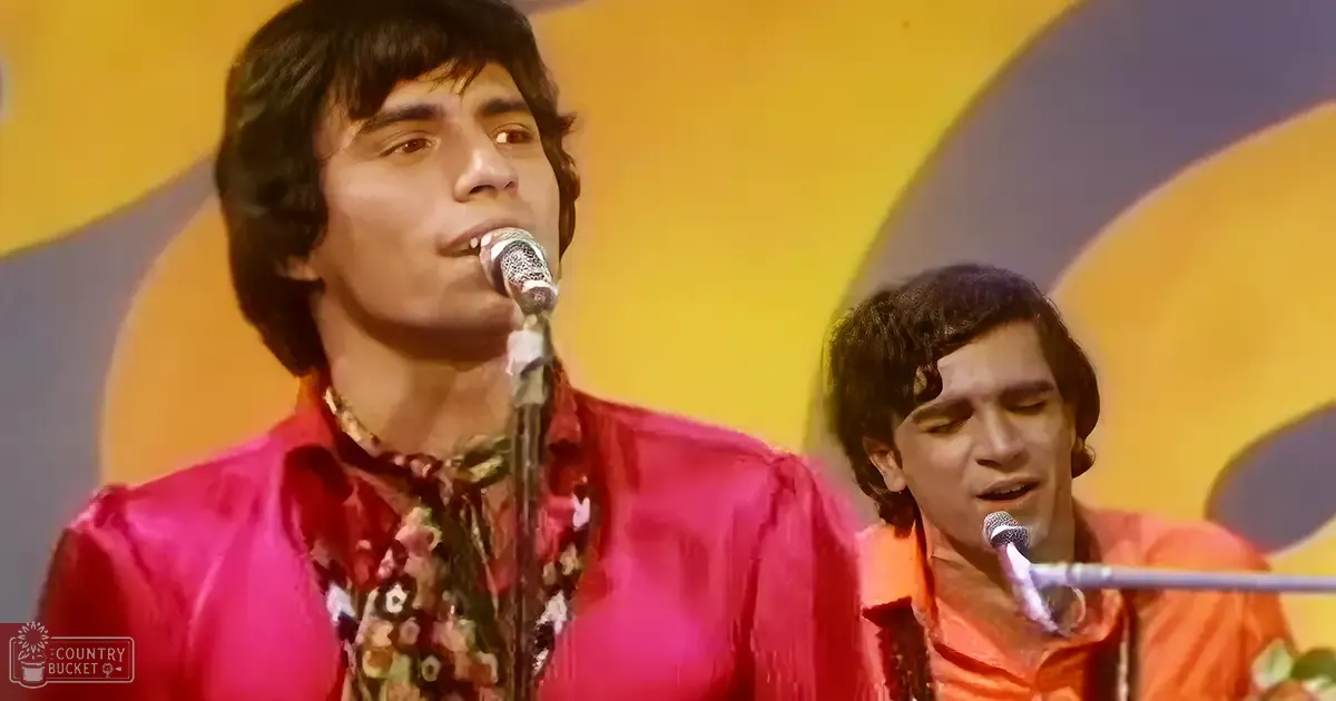 A young adult male singer wearing a bright red floral shirt performs at an indoor stage with abstract yellow, gray, and blue background.