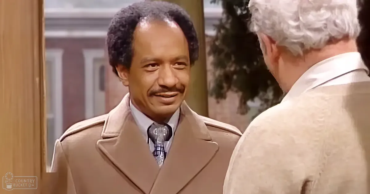 An adult male wearing a beige trench coat with a notched lapel, white collared shirt, and patterned tie stands indoors near an older adult male in a beige suit jacket. The scene includes traditional decor elements like a wooden door frame and suggests daytime lighting.