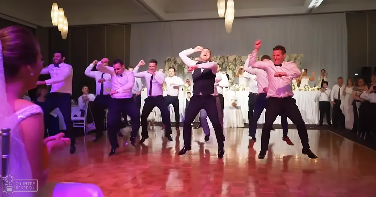 Men dancing joyfully at a wedding reception in an indoor venue with formal attire, floral arrangements, and evening lighting.