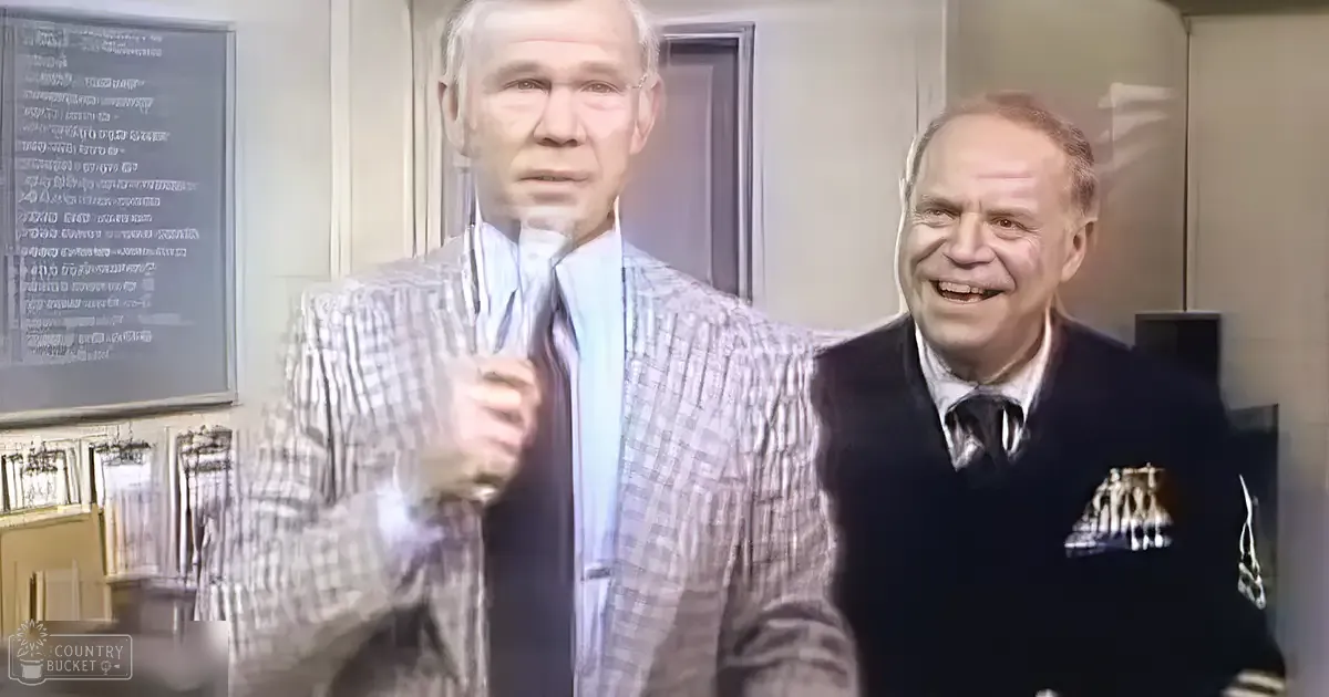 An adult male in a checkered blazer over a light blue shirt with a dark tie stands next to another adult male wearing a dark sweater vest and patterned tie. The scene is indoors, possibly an office or classroom from the mid-20th century.