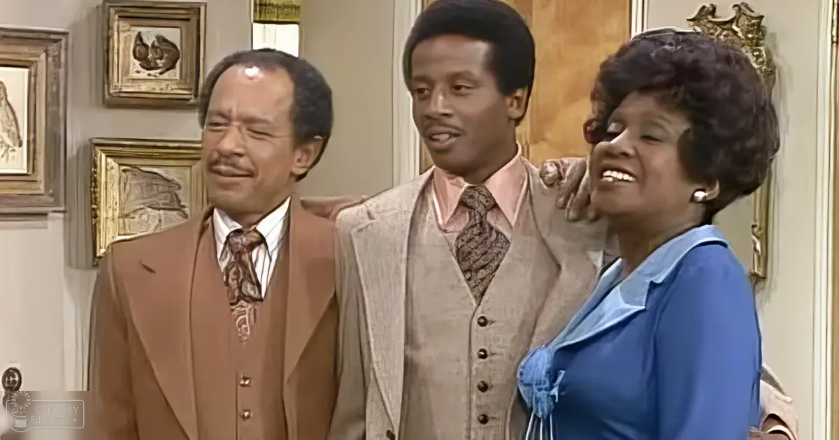 A sitcom scene set in an indoor home environment with three adults: a middle-aged man in a brown suit, another adult male in a light beige vest over a pink shirt, and an older female wearing a blue blouse and scarf. The background includes framed artwork on the wall and a wooden door.