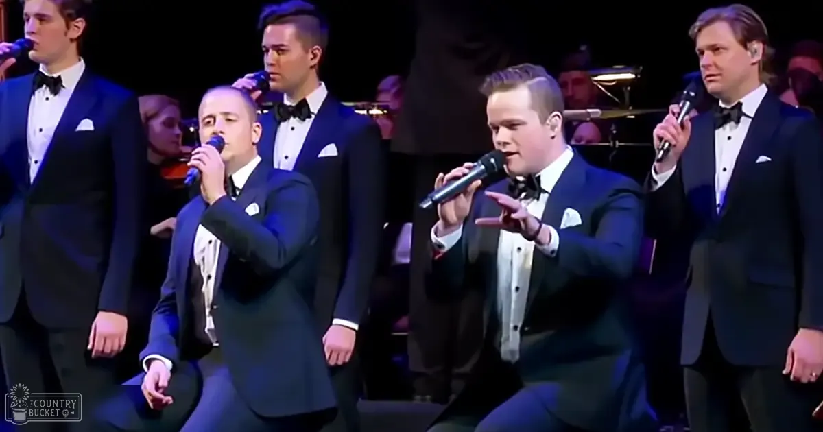 A group of adult male singers performing on a dimly lit stage, dressed in dark blue tuxedos with white shirts and black bow ties.