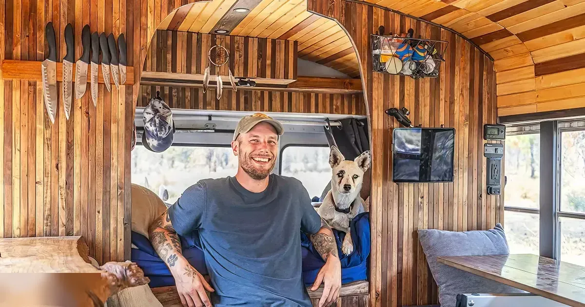 A medium-sized light brown mixed breed dog with white markings sits in a rustic RV interior, surrounded by wooden paneling and various decor items under natural daylight.