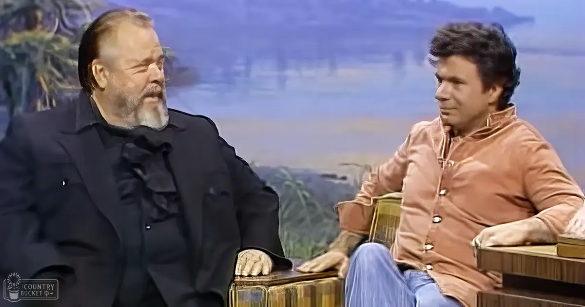 Two individuals seated in a talk show setting with simple wooden chairs and books on a small table, artistic backdrop of water and trees, soft indoor lighting, spring or summer season.