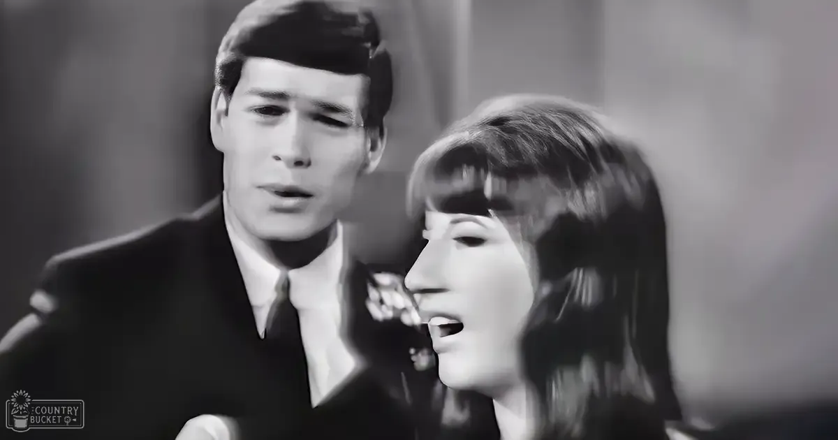 A musical performance scene with a young adult male singer wearing a dark suit from the mid-20th century and a female performer in a dark, high-necked dress on stage.