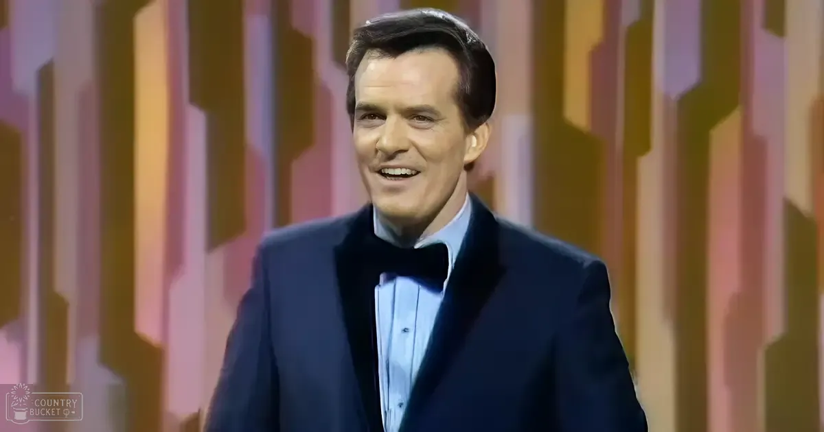 An adult male in a dark blue tuxedo with a black bow tie and light blue shirt stands against an abstract, colorful geometric backdrop during what appears to be a formal event. Text reading 'Madly Odd' is visible.