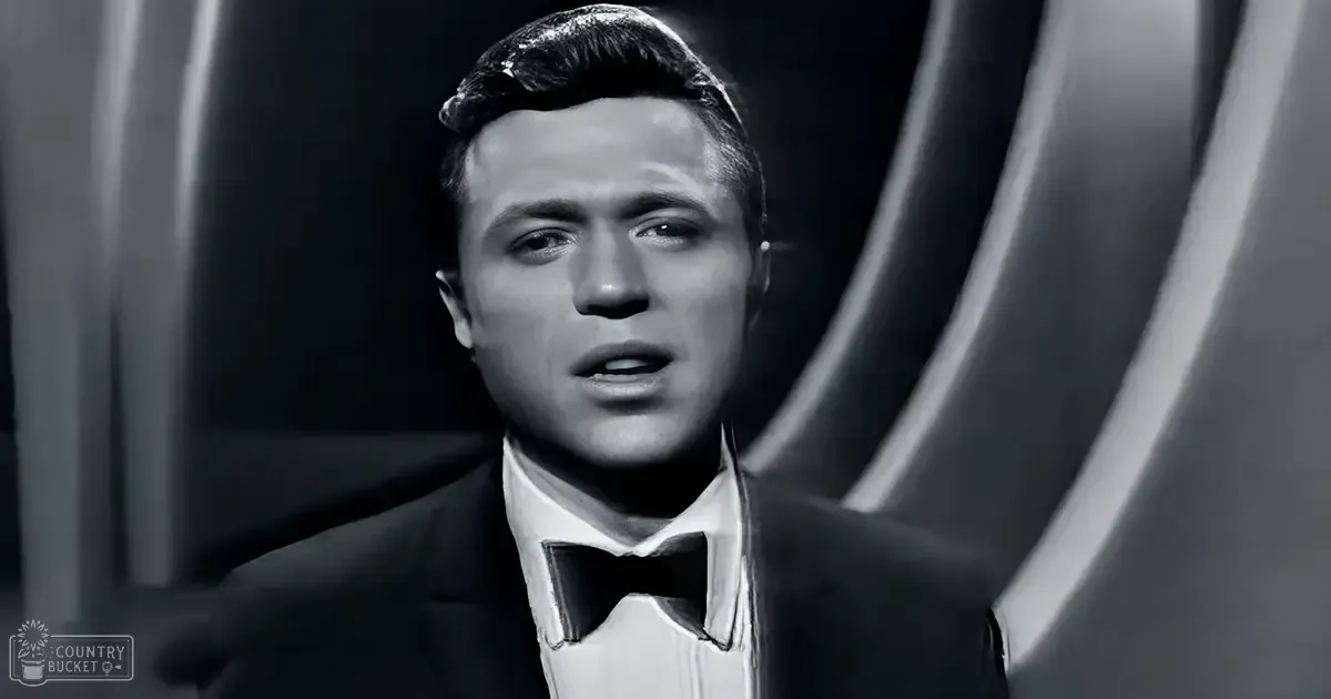 A young adult male in a formal tuxedo with a bow tie stands indoors against a dark, draped backdrop.