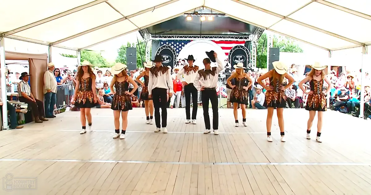 A dance performance at an outdoor event in Texas with performers wearing black and orange patterned dresses and cowboy attire, under a large tent on a sunny day.
