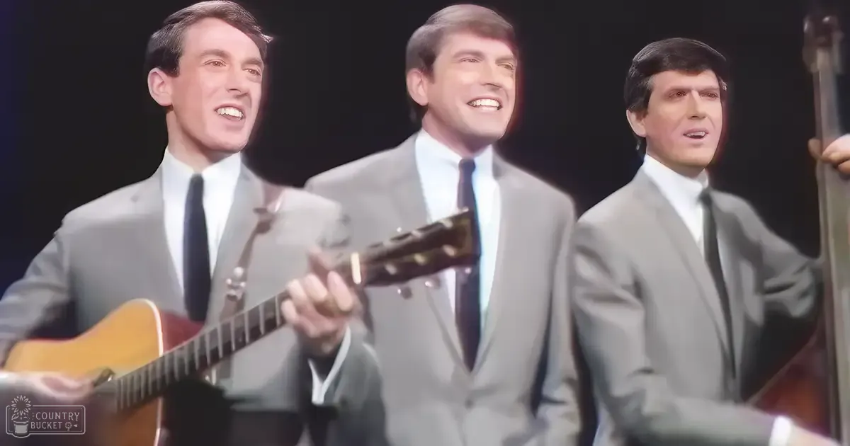 A mid-20th-century musical performance scene with three young adult male performers wearing matching light gray suits, dramatic stage lighting highlighting them against dark curtains, and musical instruments like an acoustic guitar and a double bass in the foreground.
