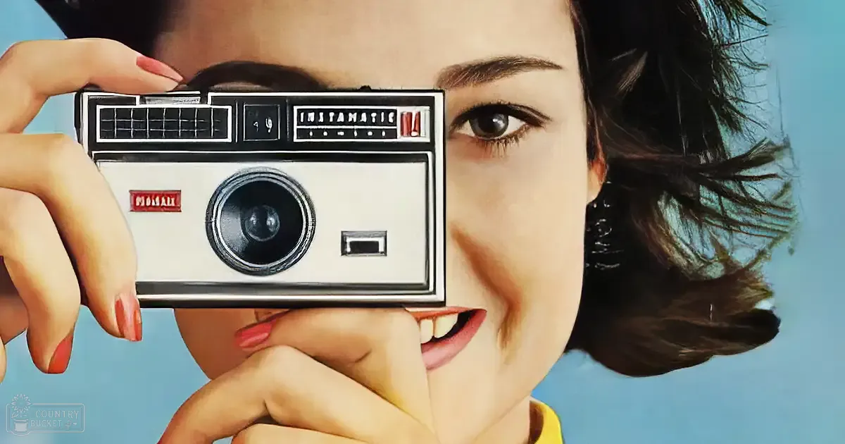 A young adult female in a mid-20th century-style yellow garment stands against a plain blue background, holding a vintage Instamatic camera with 'MOTAR' and 'INSTAMATIC' printed on it.