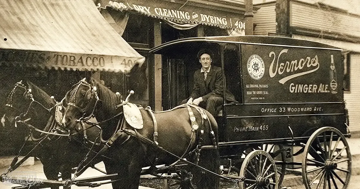 Two sturdy adult horses stand beside a wooden horse-drawn delivery wagon labeled 'Vernors Ginger Ale' in an urban setting with late 19th to early 20th-century architecture and muted sepia tones.