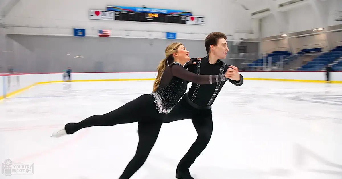 A young adult female figure skater in a black sequined leotard and male partner in a patterned black outfit skate gracefully on an indoor ice rink with banners and seating areas visible in the background.