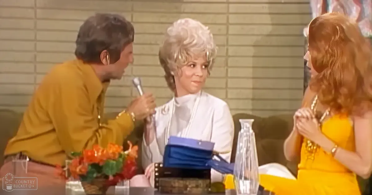 Three individuals in a talk show setting with mustard-yellow shirts and accessories. Furniture includes a dark green couch and a small table with flowers.